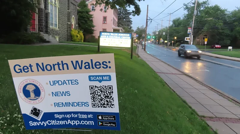 Trees line both sides of Main Street in North Wales, as a sign directs residents to sign up for the town’s “Savvy Citizen” notification alerts, in summer 2023.