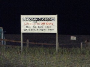 As a Coast Guard boat searches for a missing swimmer nearby on Sunday night, a sign at Ninth Street Beach warns visitors to stay out of the water after the lifeguards leave for the day.