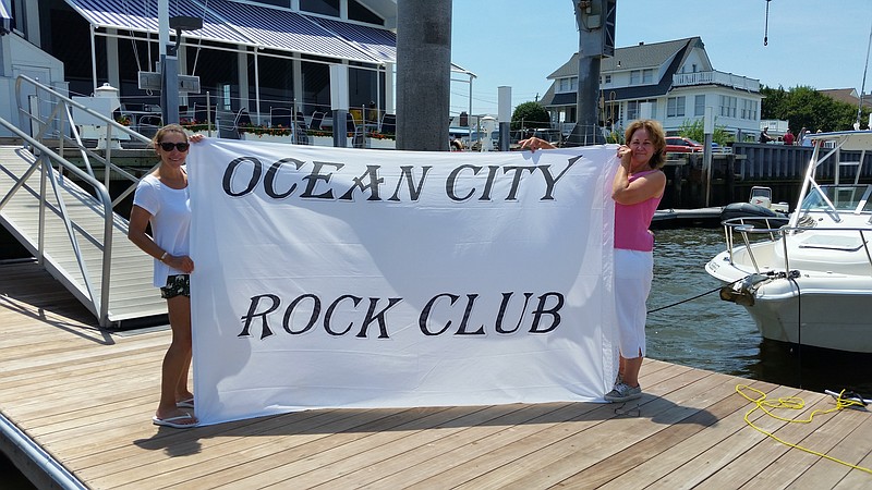 At the Ocean City Yacht Club, member Faith Camp-O'Donnell, left, and Kathryn Doms, who serves as rear commodore, unfurl an "Ocean City Rock Club" banner that will be on display for the rock-themed Night in Venice celebration.