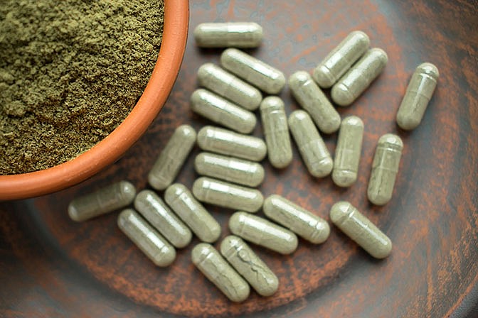 Supplement kratom green capsules and powder on brown plate. Herbal product alt-medicine kratom is  opioid. Home alternative pain remedy, opioid addiction, dangerous painkiller, overdose. Close up. Selective focus