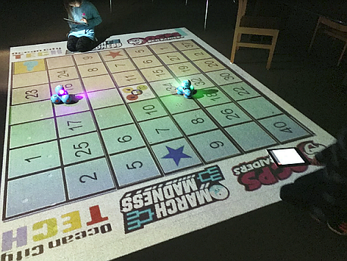 3rd grade student, Isabelle Tolbert, creating an algorithm for her robot to move towards the higher points on the board. The customized game board is projected onto the floor using a smart projector. As the robot moves and stops in the numbered squares the points disappear from the board.