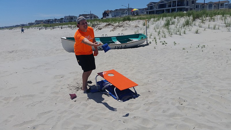 McElroy tosses a bean bag while standing in front of a beach chair using the cornhole game attachment.