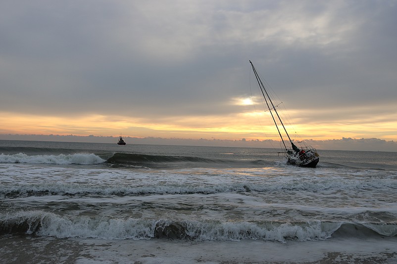 The sailboat is pulled back into the ocean by the towing vessel seen in the distance to the left.