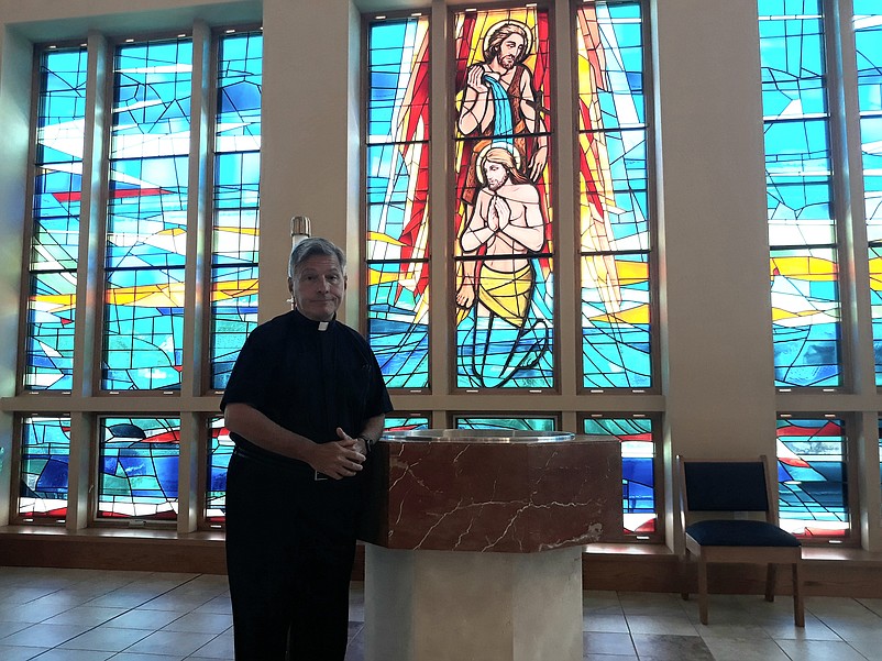 Father Perry stands in front of a series of stained glass windows in the church depicting the baptism of Jesus.