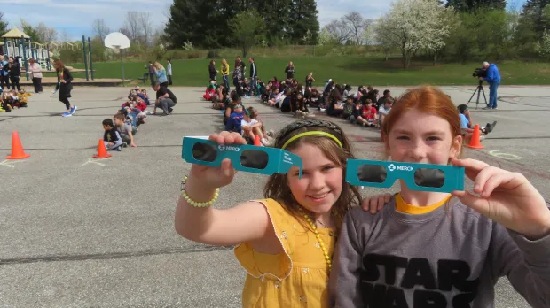 2 girls hold solar eclipse viewing glasses outdoors with other students in the background.