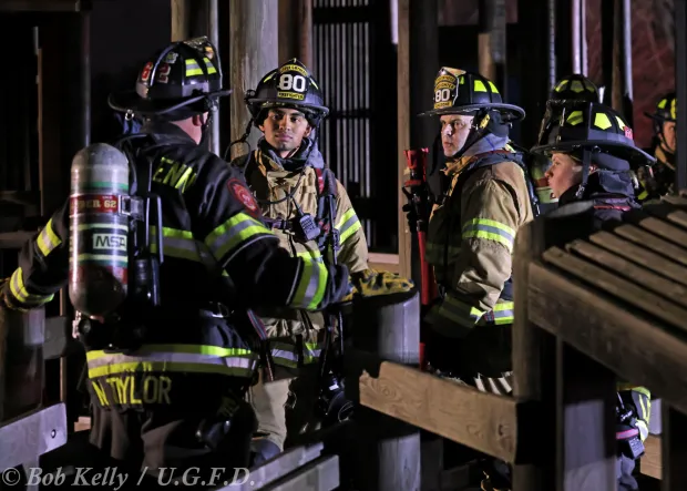 Upper Gwynedd Fire Department firefighters listen to one of their counterparts from the North Penn Volunteer Fire Company firefighters at a fire scene. (Photo by Bob Kelly for UGFD)
