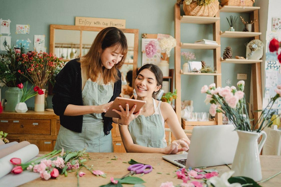 Free Content multiethnic female florists in aprons at table with laptop and browsing tablet while preparing for order in creative floristry studio Stock Photo