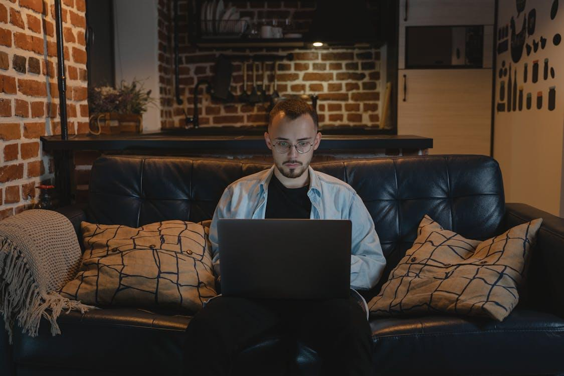 Free Man working remotely on a laptop in a cozy, stylish apartment with exposed brick walls and modern decor. Stock Photo