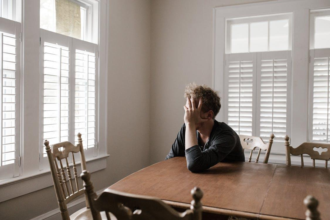 Free A man sits alone at a table in a bright room, displaying deep contemplation. Stock Photo