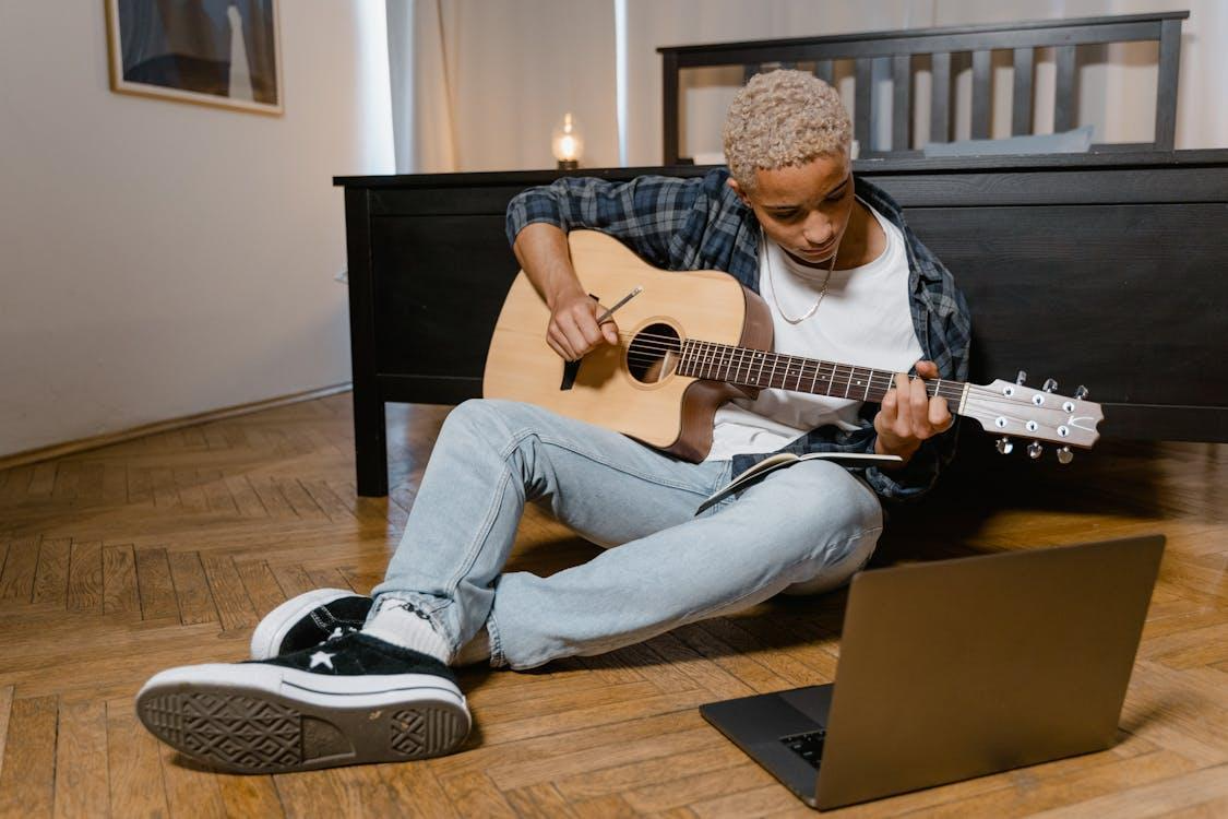 Free Teen sitting on the floor, playing acoustic guitar beside a laptop, creating music at home. Stock Photo